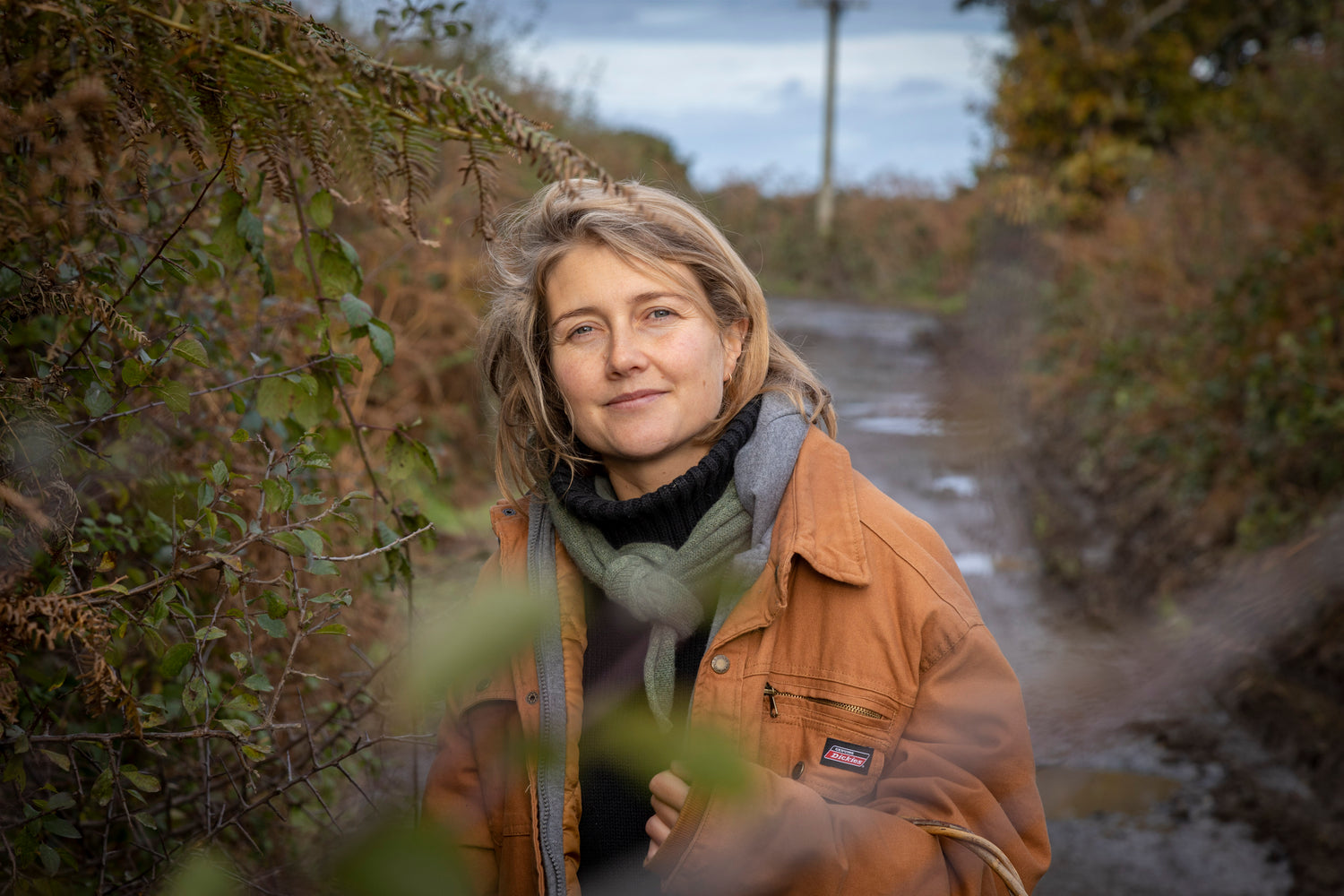 Emily Archer wearing an orange jacket and grey scarf. She is surrounded by foliage and is on a dirt road.